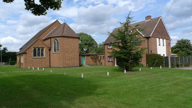 A church with a brick facade and a smaller adjacent building, surrounded by a grassy field and trees.