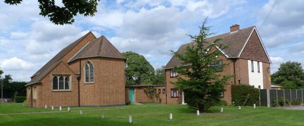 A church with a brick facade and a smaller adjacent building, surrounded by a grassy field and trees.