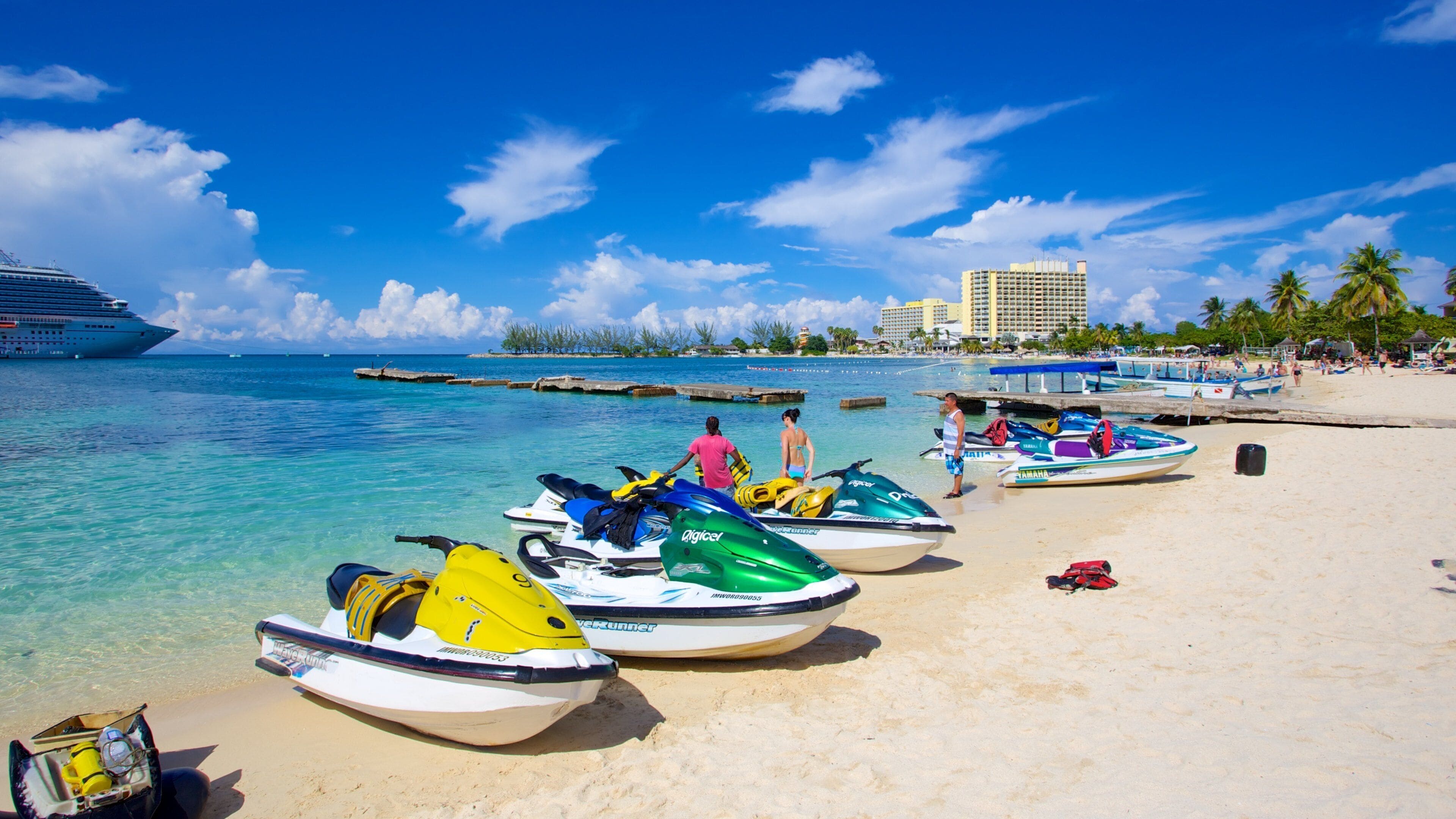 Uma praia com jet skis, um cruzeiro e um resort ao fundo.