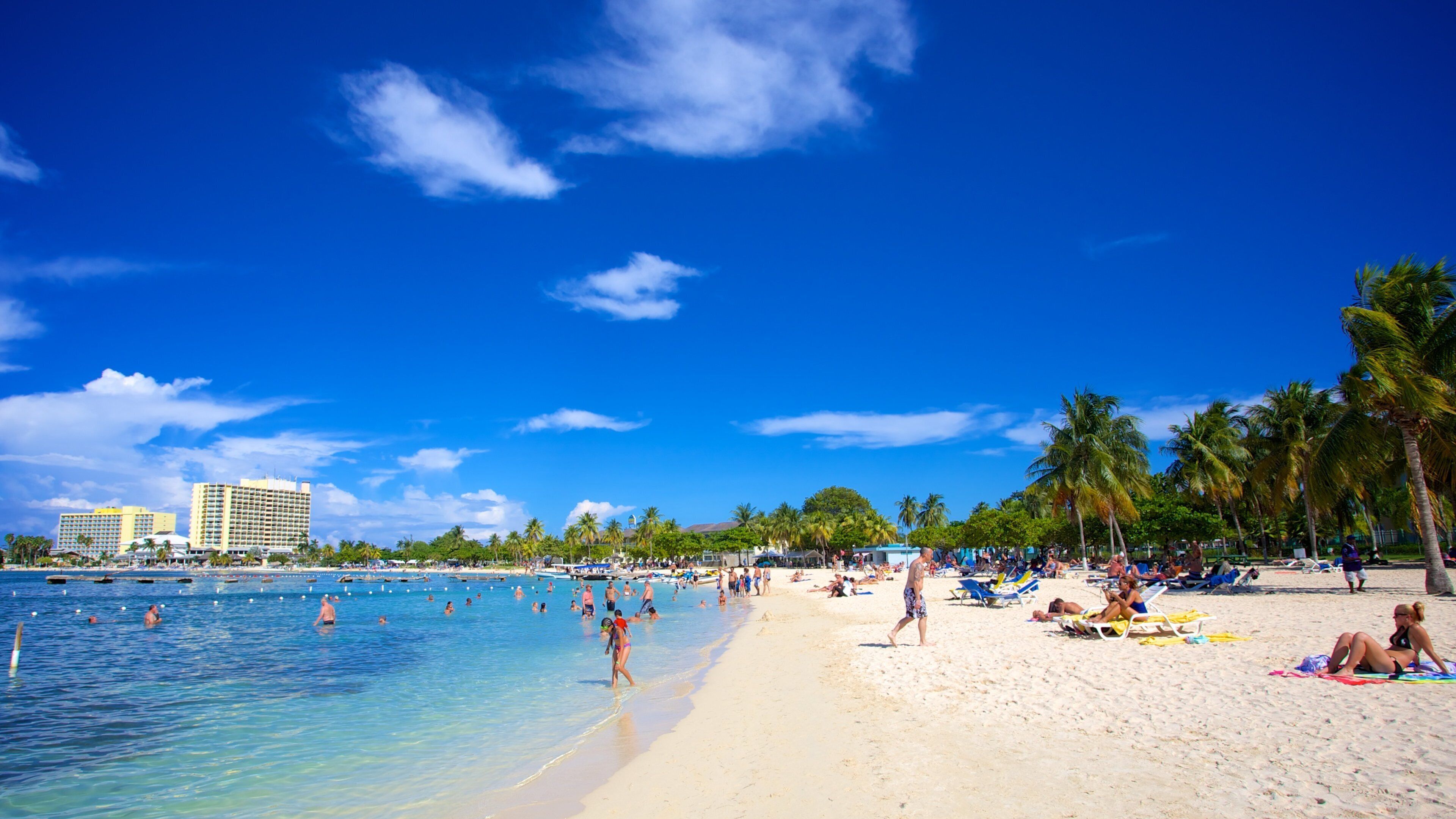 A beach with clear blue water, palm trees, and a hotel in the background.