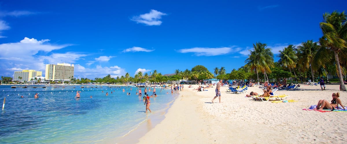 A beach with clear blue water, palm trees, and a hotel in the background.