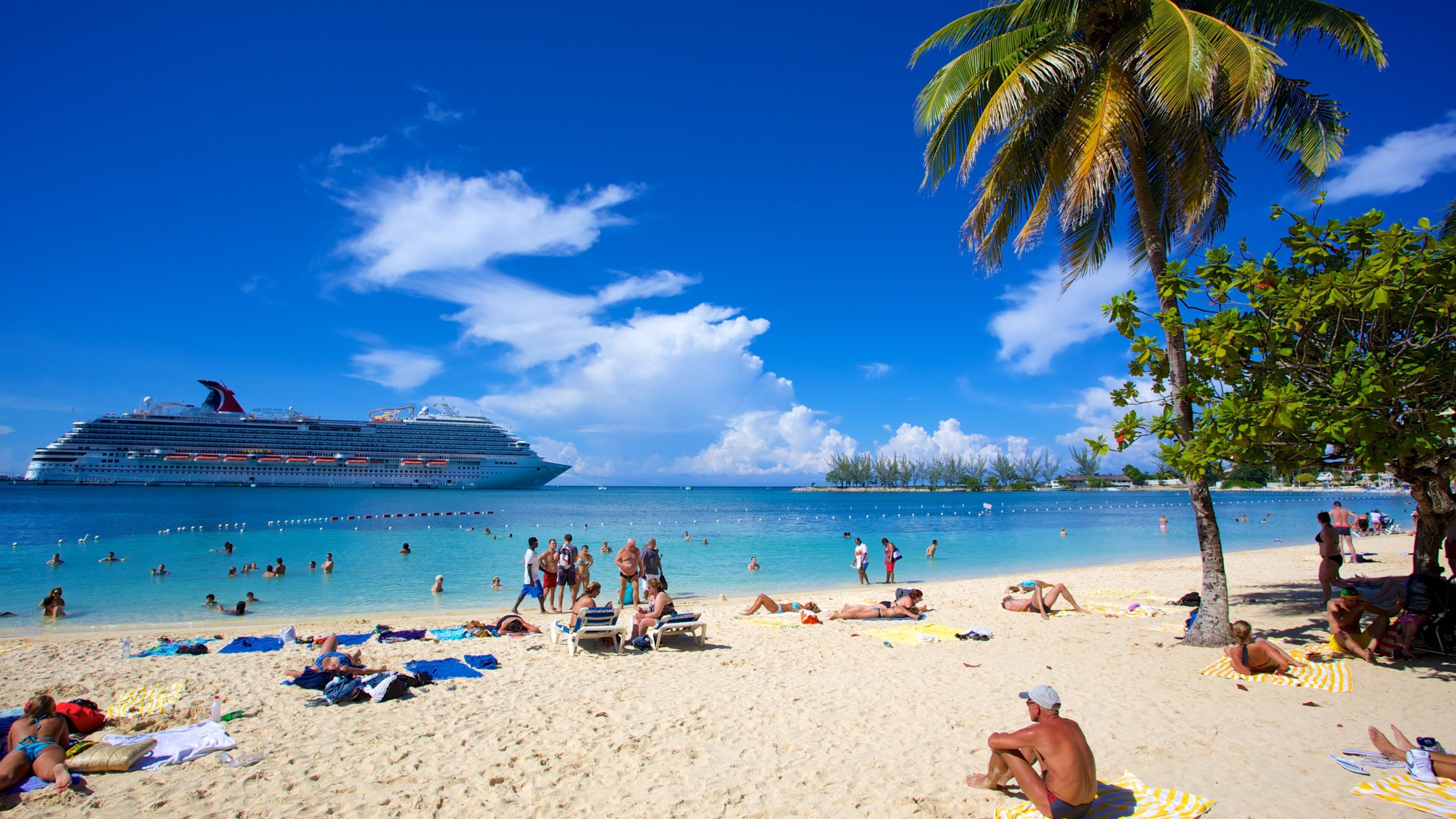 Una playa con gente tomando el sol, un crucero al fondo y un cielo azul despejado.