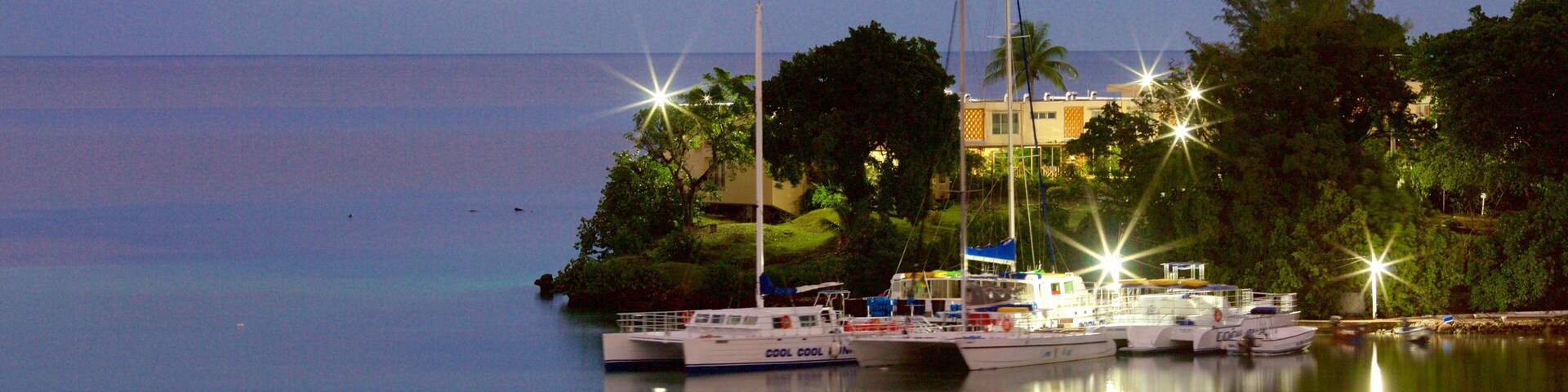Um porto ao anoitecer, com barcos atracados, um farol e um edifício rodeado de árvores.