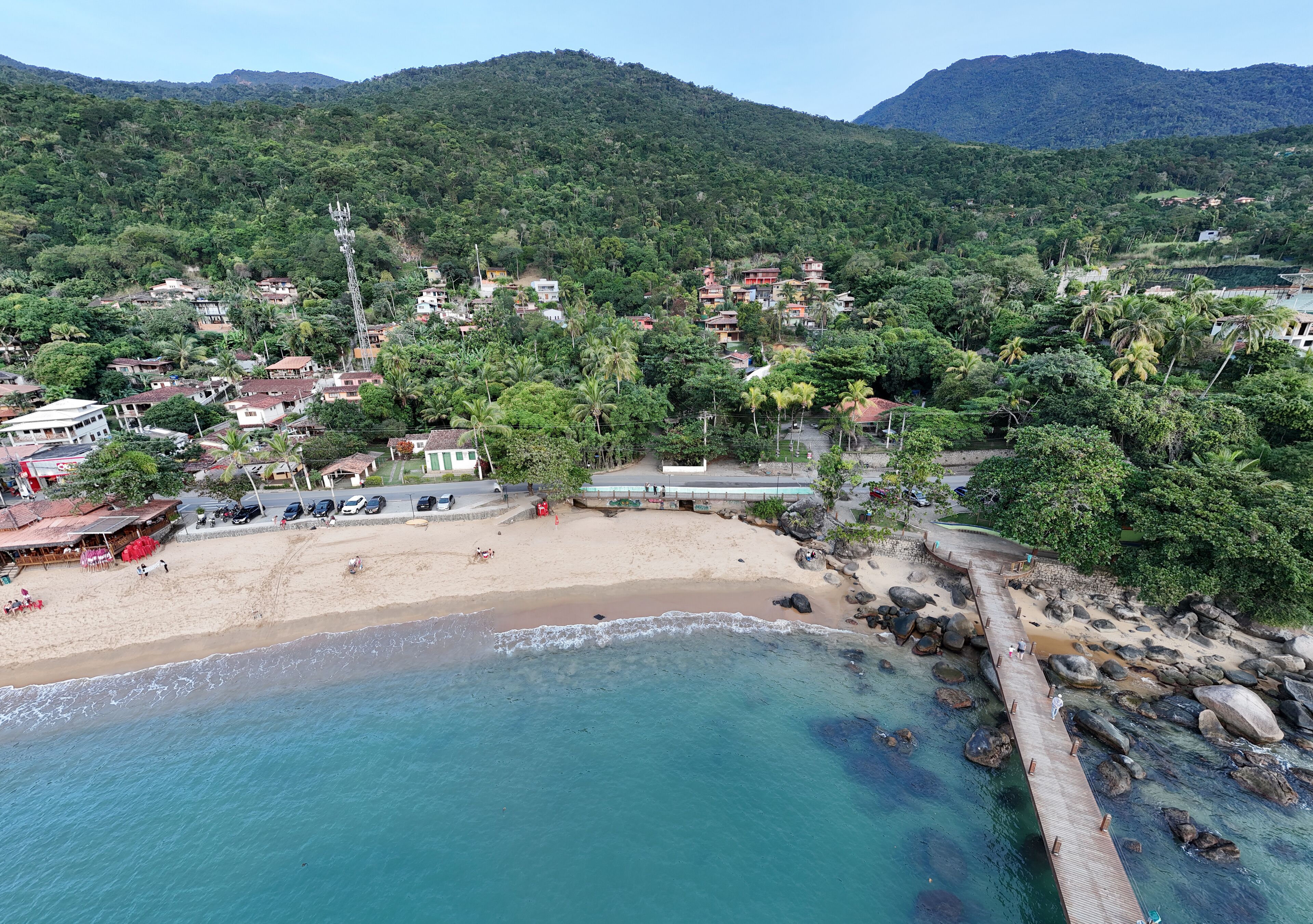 Visão aérea do píer na Praia Grande em Ilhabela, SP, com mar azul, faixa de areia e estrutura de madeira sobre a água, em cenário litorâneo tropical