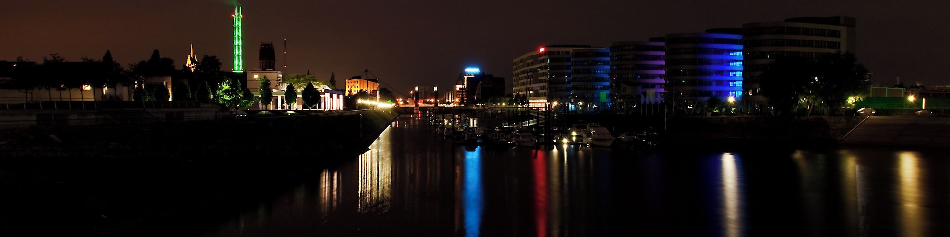 Une vue nocturne d’une ville, avec des bâtiments illuminés qui se reflètent dans une étendue d’eau.