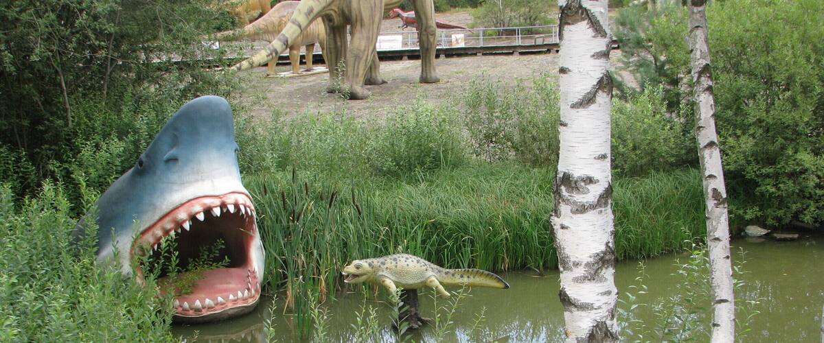 A crocodile near a birch tree and a large shark sculpture in a pond with dinosaurs in the background.