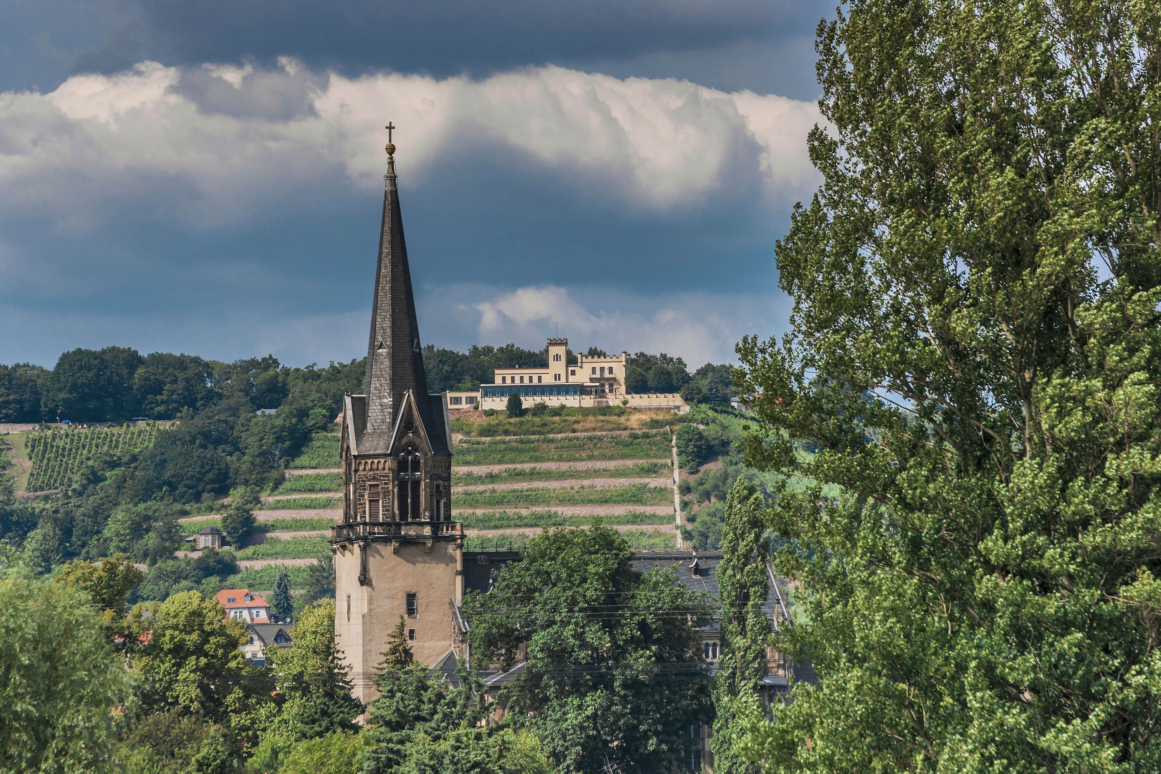 A church tower, a castle, and vineyards on a hillside under a cloudy sky.