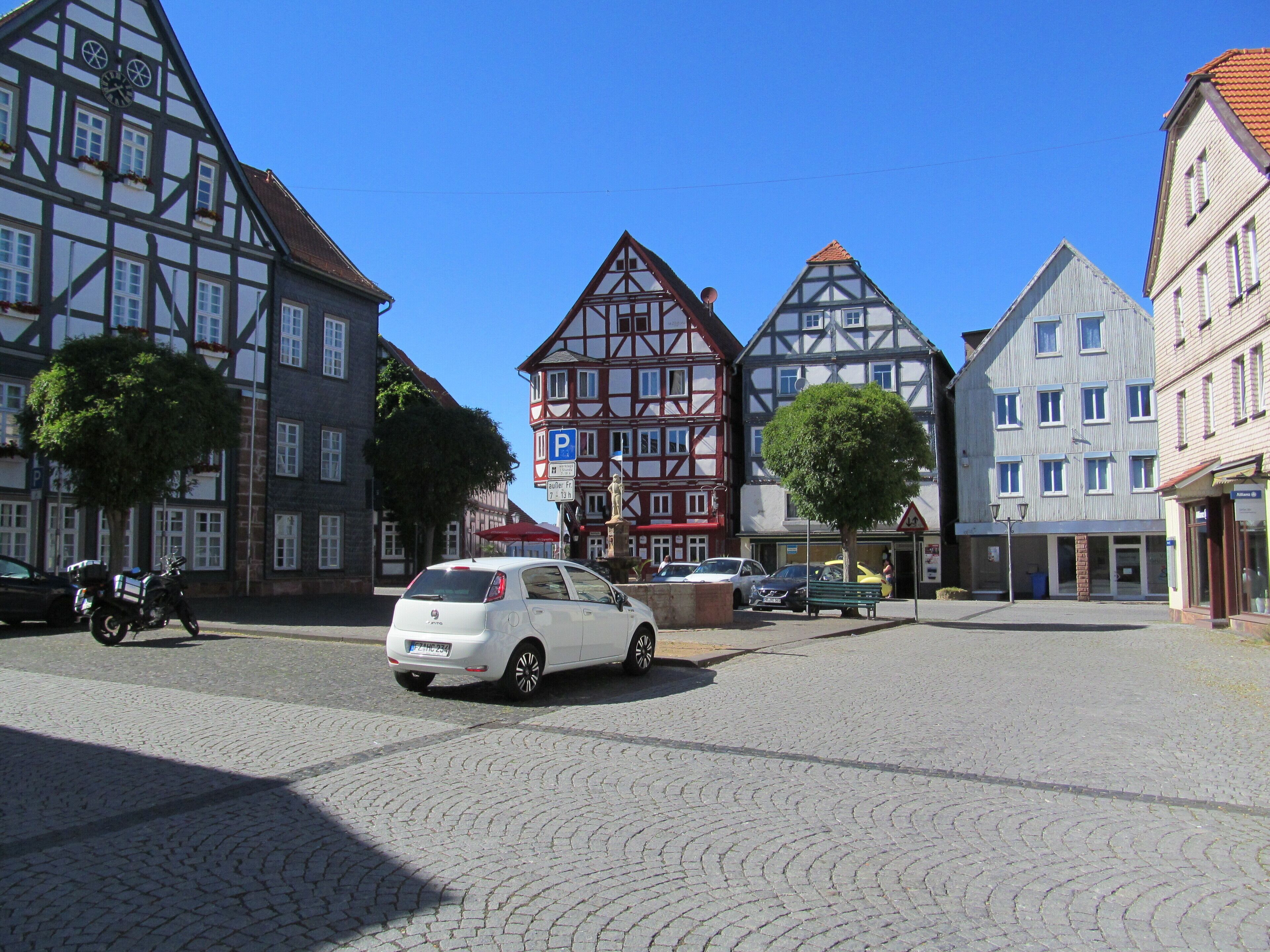 A cobblestone square with historic half-timbered buildings, a white car, and a motorcycle.