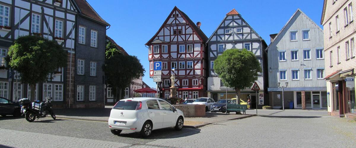 A cobblestone square with historic half-timbered buildings, a white car, and a motorcycle.