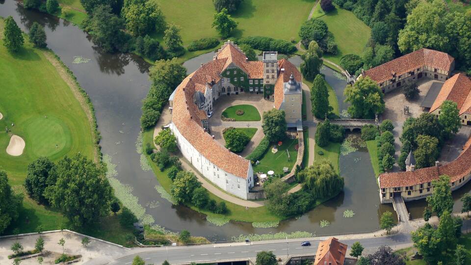 An aerial view of a castle complex with a moat, surrounded by lush greenery and a golf course.