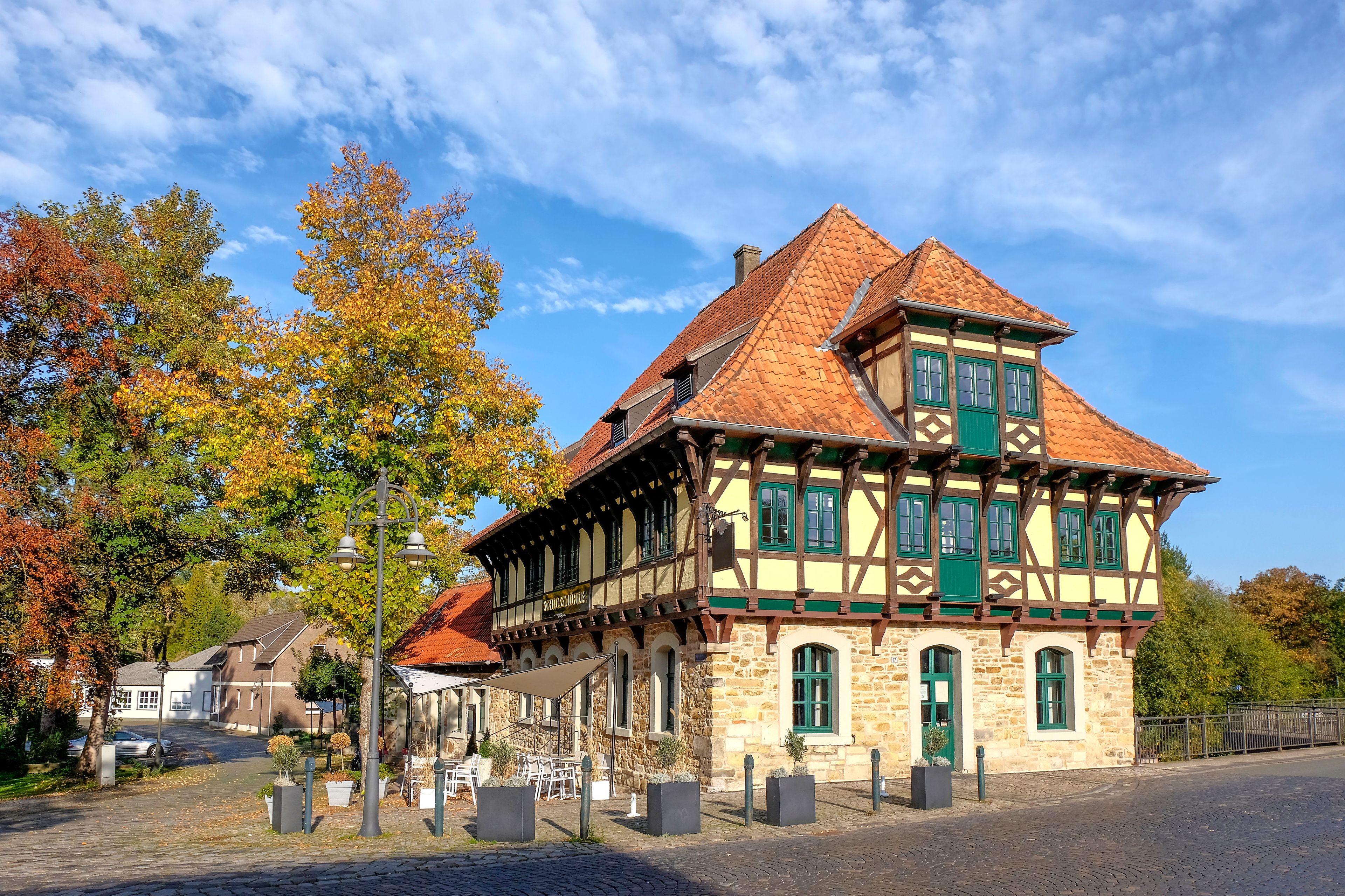 A traditional half-timbered building with a red-tiled roof, surrounded by autumn-colored trees and a cobblestone street.