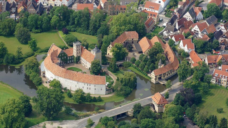 An aerial view of a historic town with a prominent castle, river, and surrounding greenery.