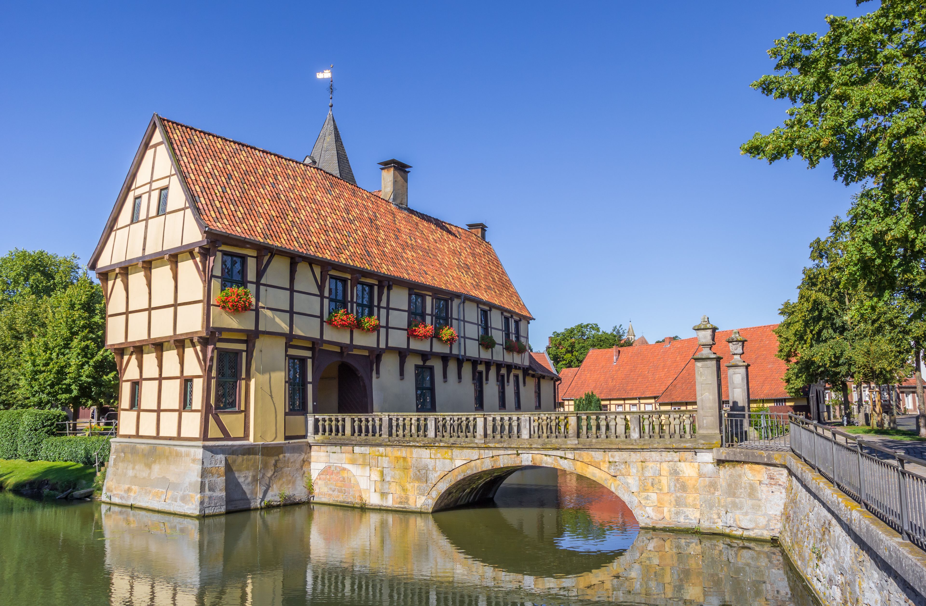 A traditional half-timbered house with a red-tiled roof, a stone bridge, and a calm river reflecting the building.