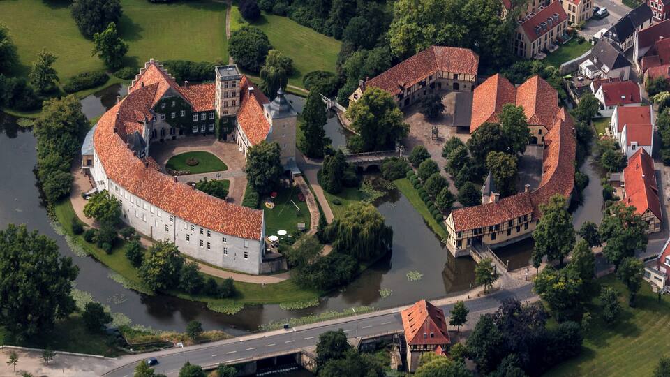 An aerial view of a historic castle complex with red-tiled roofs, surrounded by lush greenery and a river.