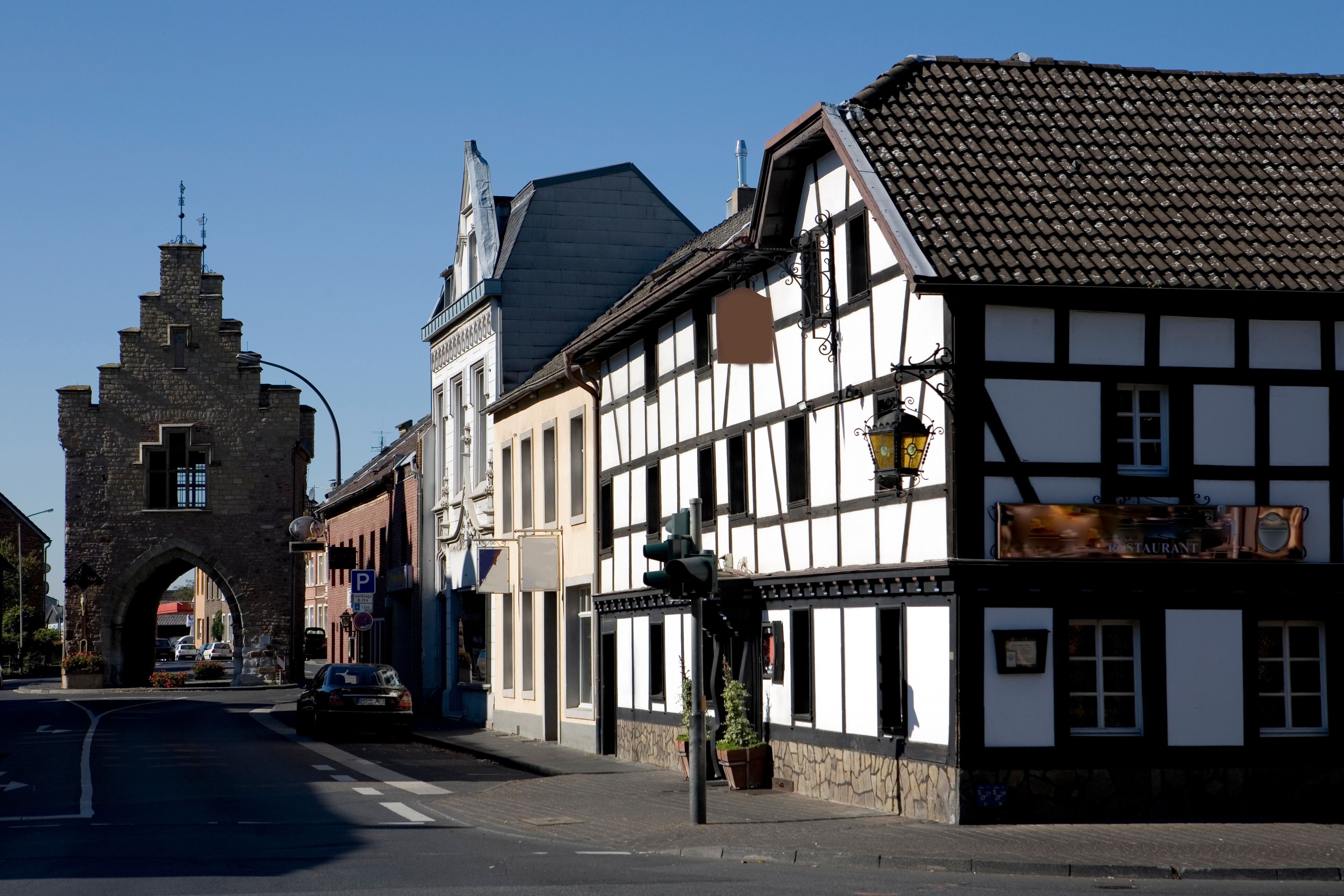 A historic street with a stone archway, traditional half-timbered buildings, and a restaurant sign.