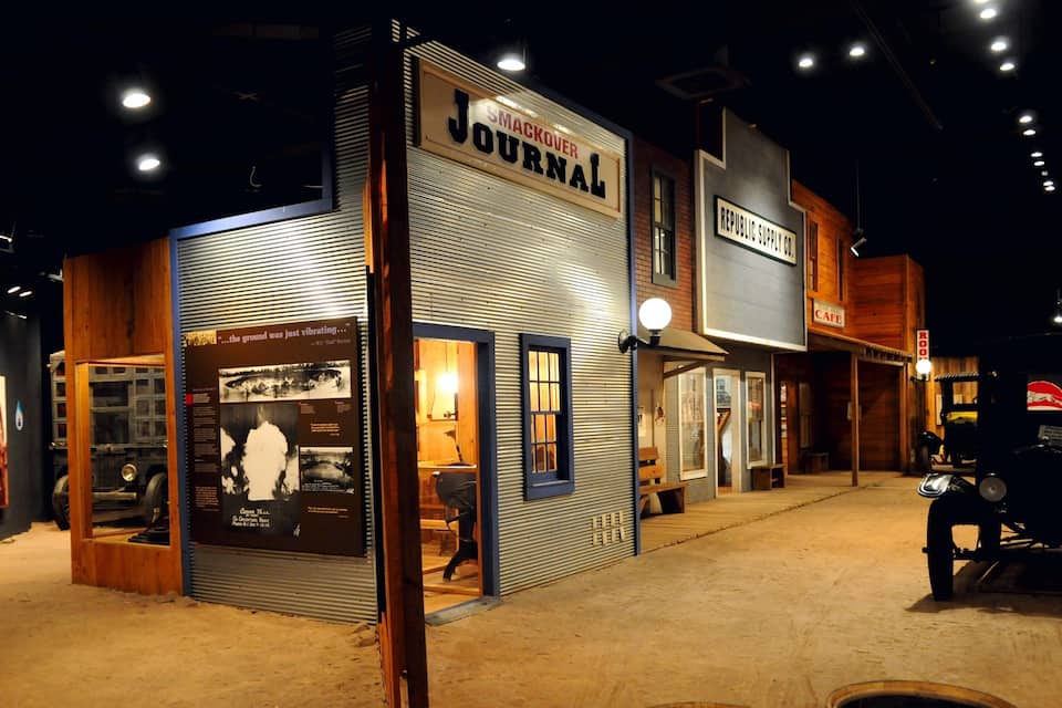 A museum exhibit with a 'Smackover Journal' sign, a 'Republic Supply Co.' sign, and a vintage car display.