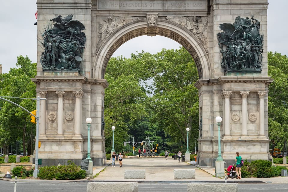 Un arco de piedra ornamentado con esculturas en relieve a ambos lados, rodeado de frondosos árboles y elementos urbanos.