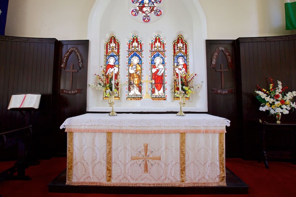 A church altar with a white cloth and a cross, surrounded by stained glass windows and floral arrangements.