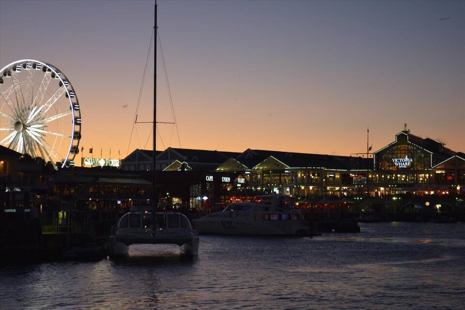 En havnefront med et pariserhjul, en båd og oplyste bygninger, herunder Victoria Wharf.