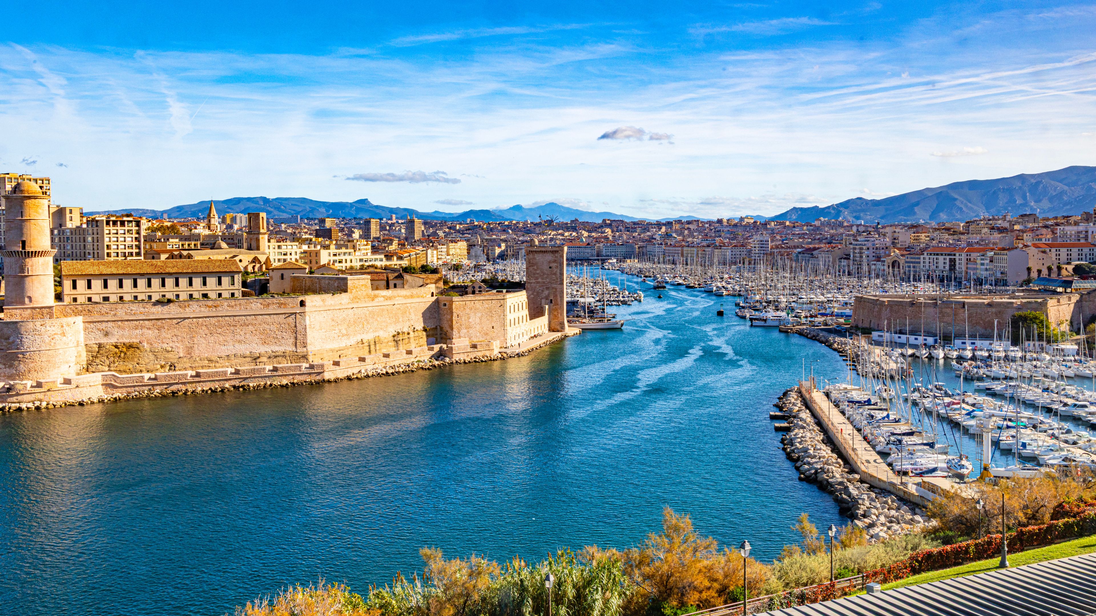 A coastal city with a historic fort, a marina with numerous boats, and a backdrop of mountains.