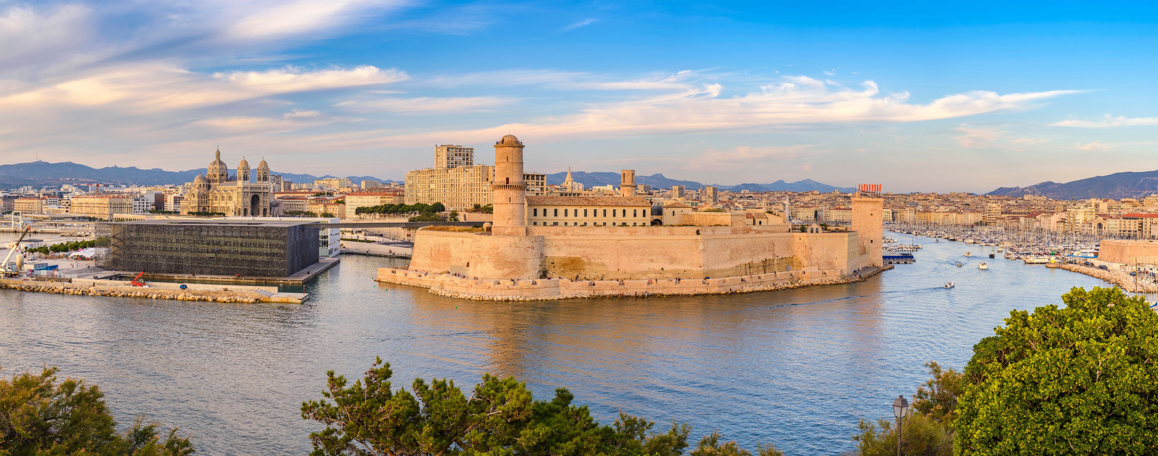 A coastal cityscape with a prominent fortress-like structure, a cathedral with domes, and a harbor with boats.