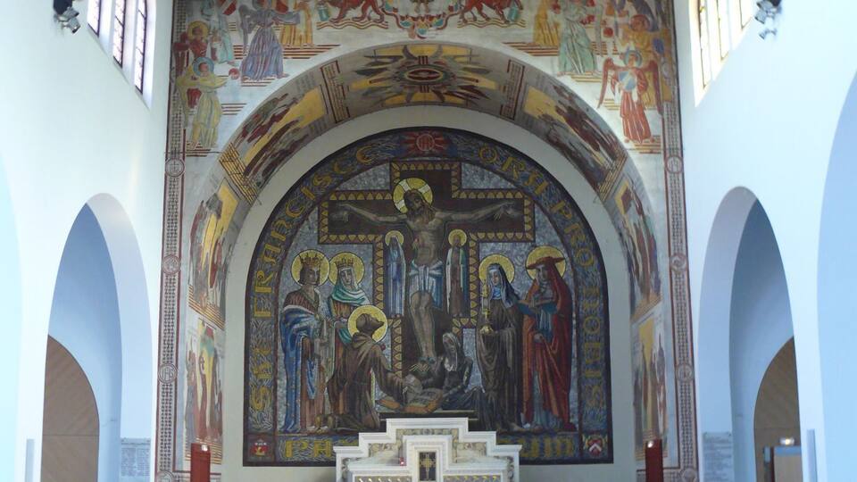 An interior of a church with red pews, a central mosaic depicting religious figures, and arched windows.