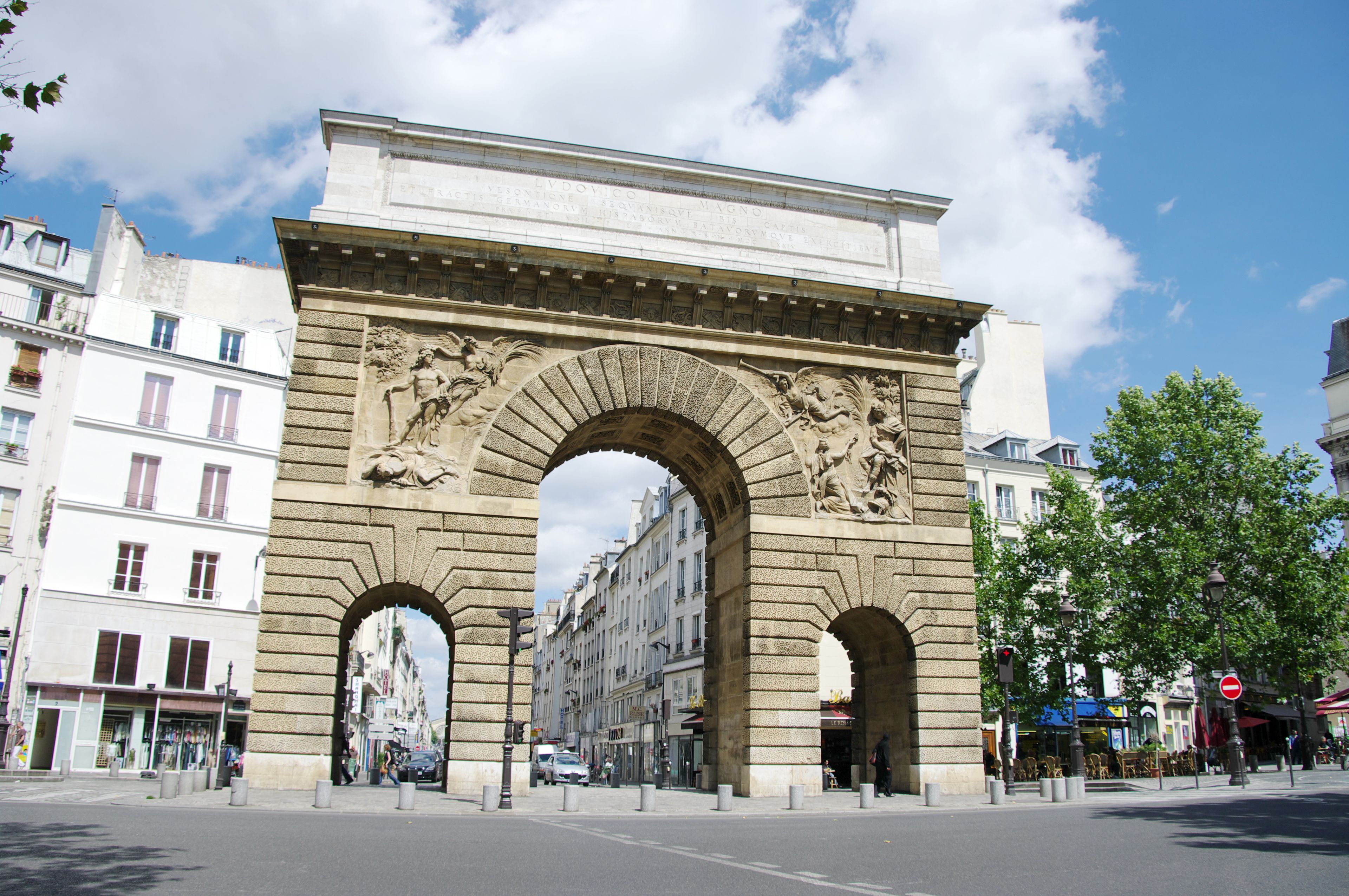 A large stone archway with intricate carvings and inscriptions, surrounded by urban buildings and a clear sky.