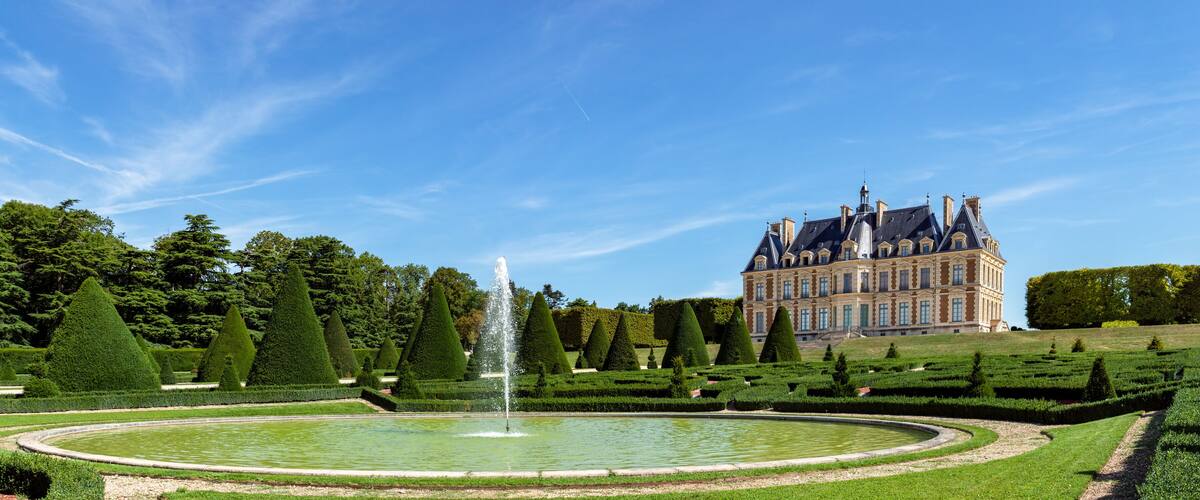 A grand château with a formal garden, including a circular pond and topiary trees.