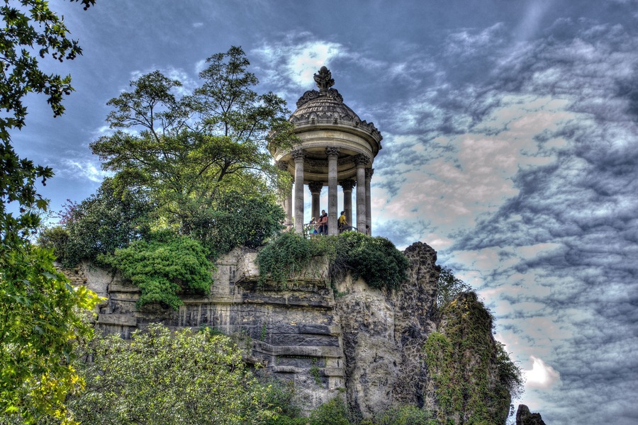 A historical stone structure with columns and a dome, surrounded by lush greenery and a dramatic sky.