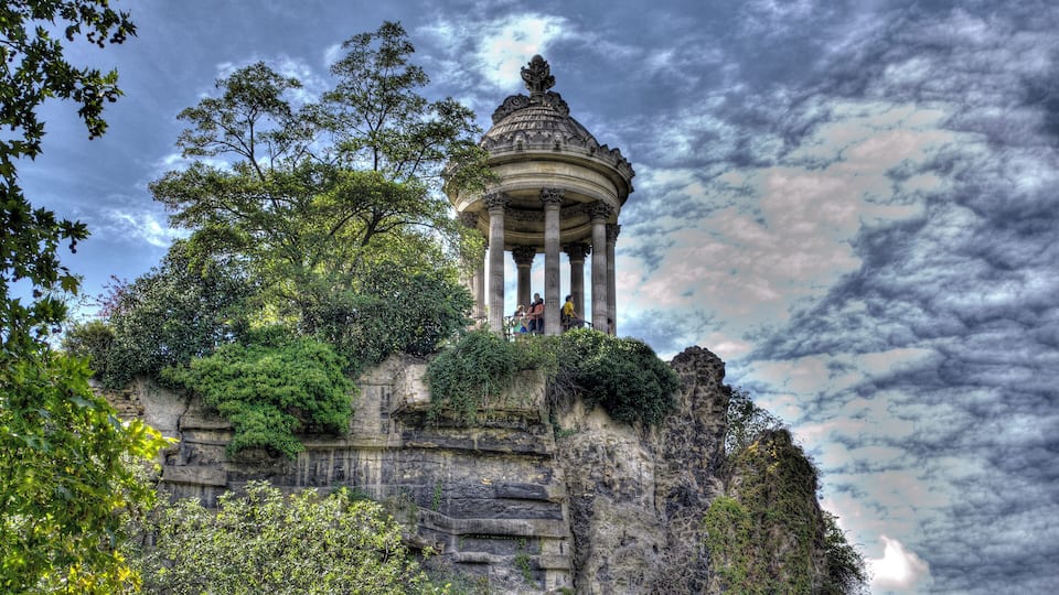 Uma construção histórica de pedra com colunas e cúpula, rodeada por vegetação exuberante e um céu dramático.