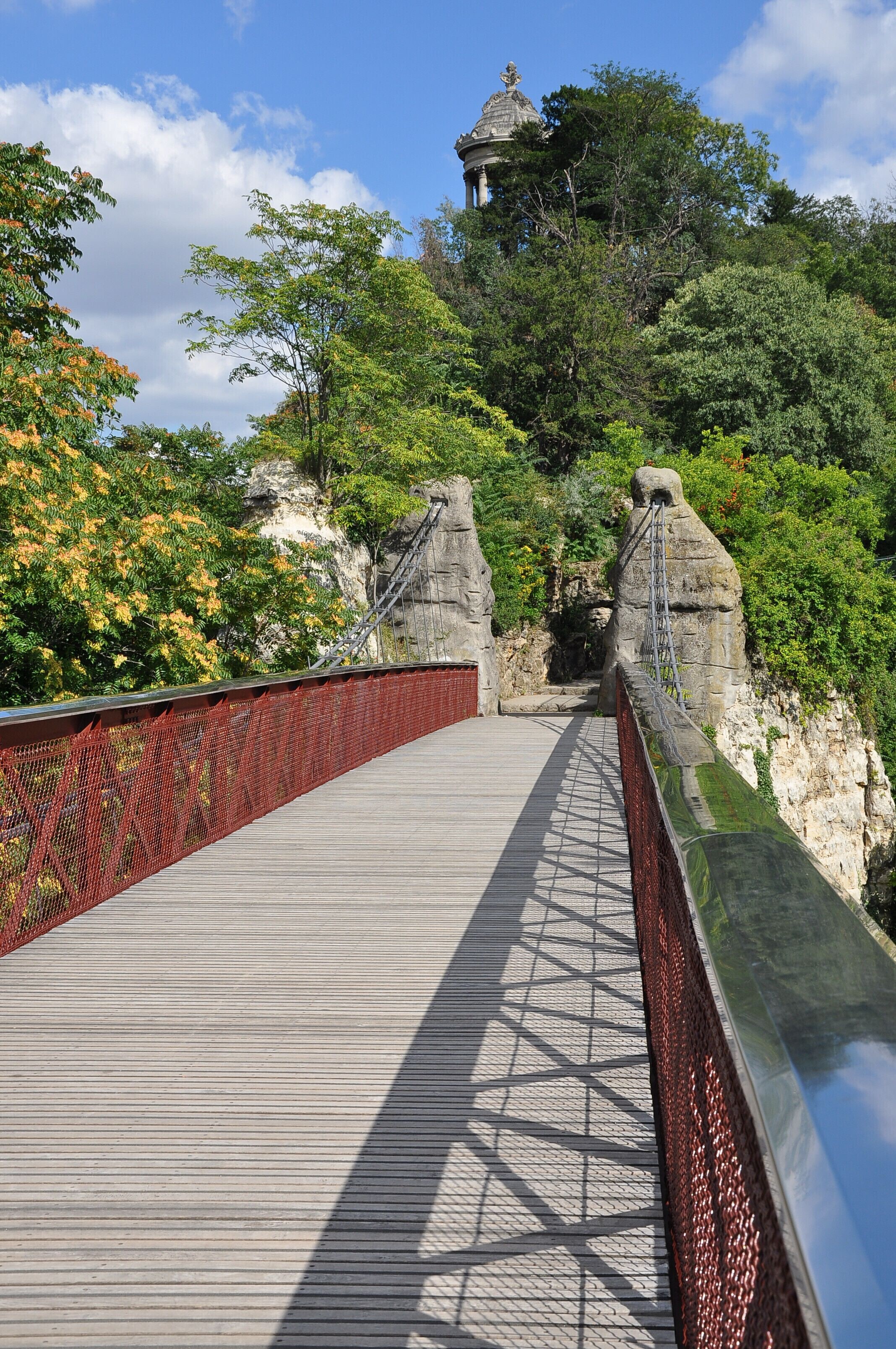 Uma ponte de madeira com grades vermelhas, rodeada por vegetação exuberante e penhascos rochosos.