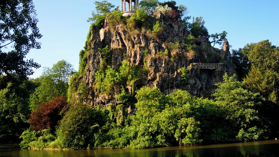 Formação rochosa com um coreto no topo, cercada por vegetação exuberante e um lago tranquilo.