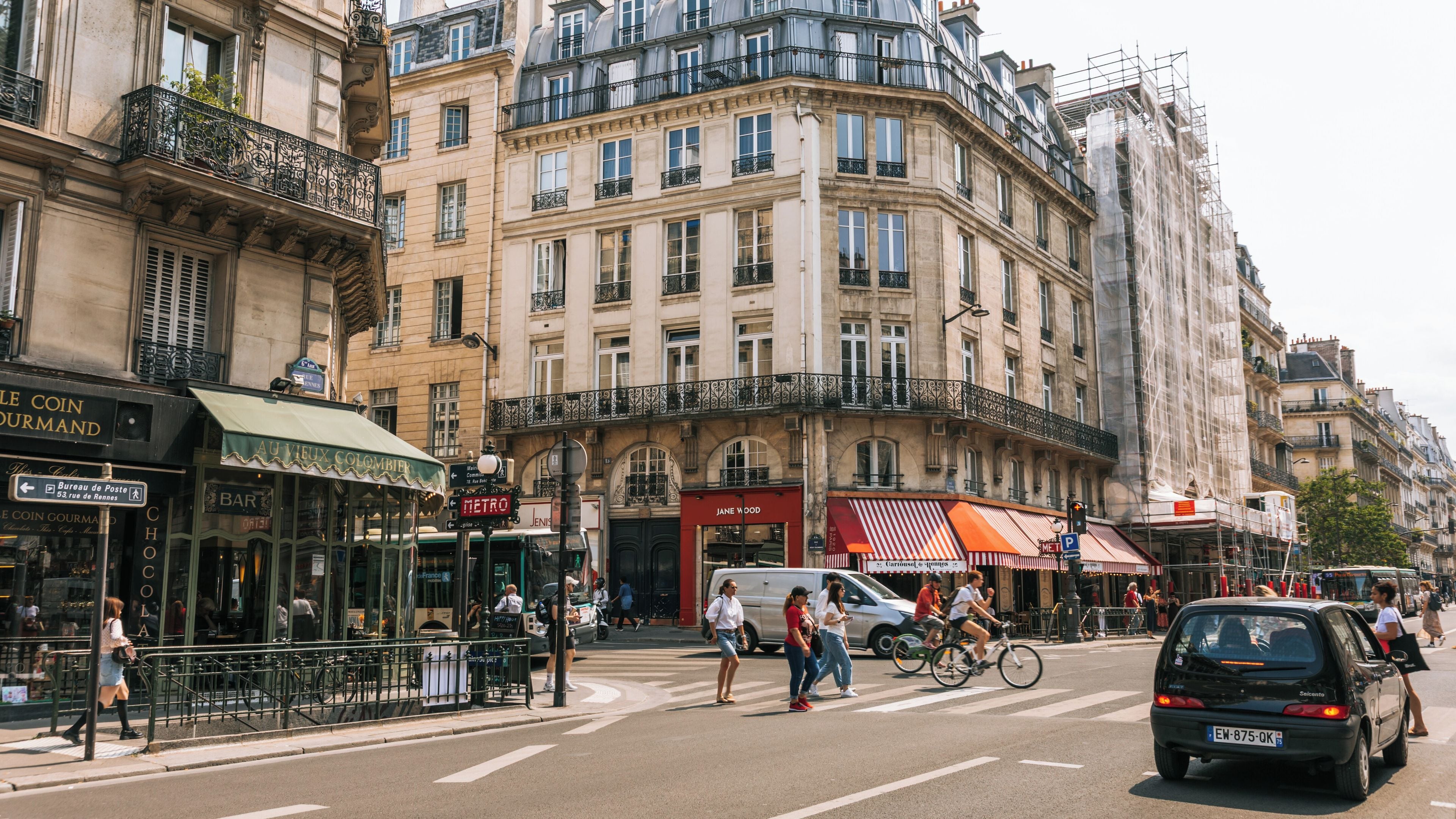 A busy urban street with pedestrians, cyclists, and vehicles. Notable buildings include a café with a green awning and a store with a red awning.