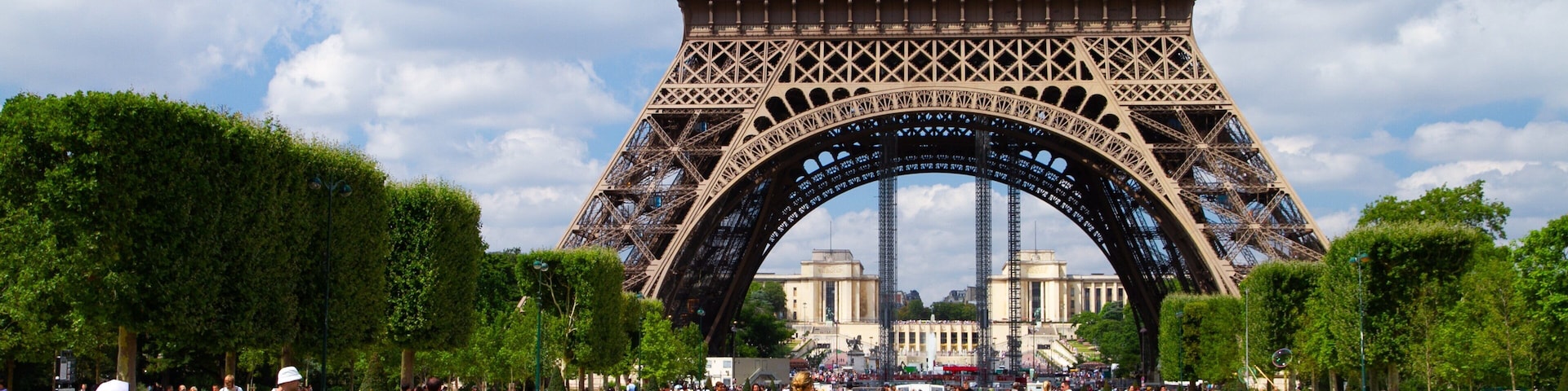 A Torre Eiffel com pessoas caminhando e sentadas na grama em primeiro plano.