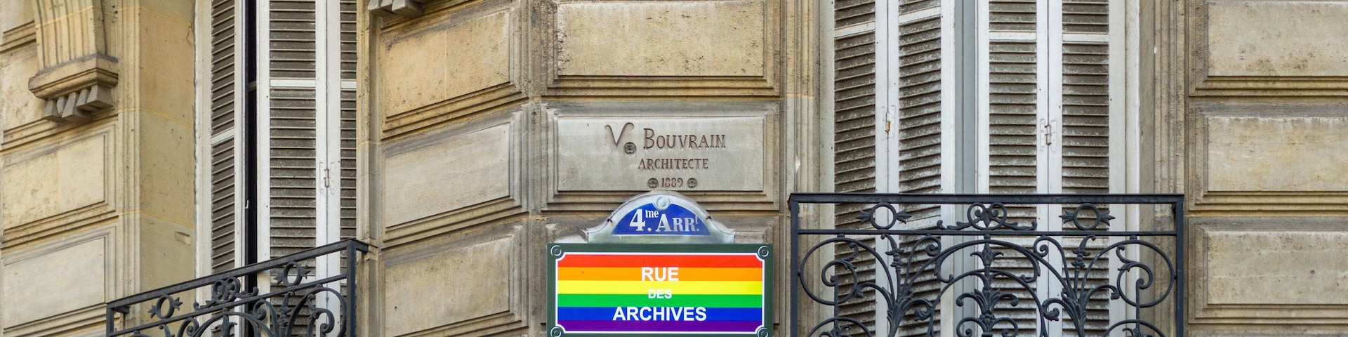Eine Gebäudefassade mit aufwendigen Eisengeländern, einer Tafel mit der Aufschrift 'Boulevard des Archives' und einem farbenfrohen Schild.