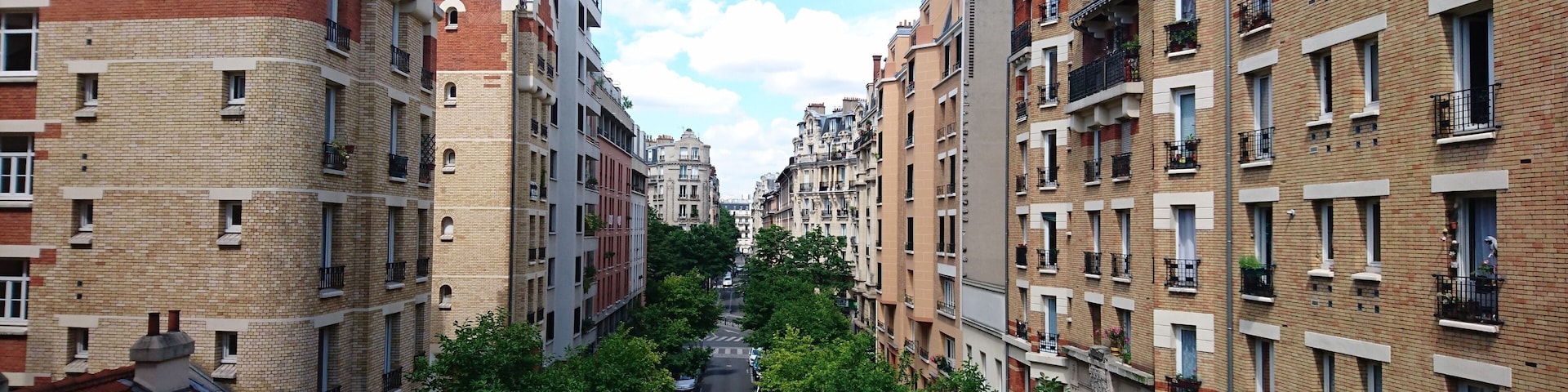 Une rue bordĂ©e dâarbres, de part et dâautre, se dressent des immeubles Ă plusieurs Ă©tages, dotĂ©s de balcons et surmontĂ©s de toits recouverts de tuiles rouges.