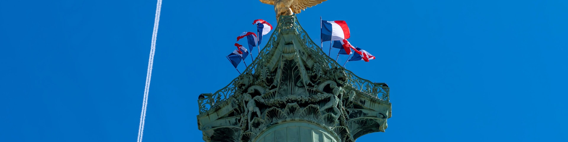 Paris, France - 04 25 2026: Place de la Bastille. Detail View of the genius of Liberty golden statue on July column, national French flags around with sunny blue sky
