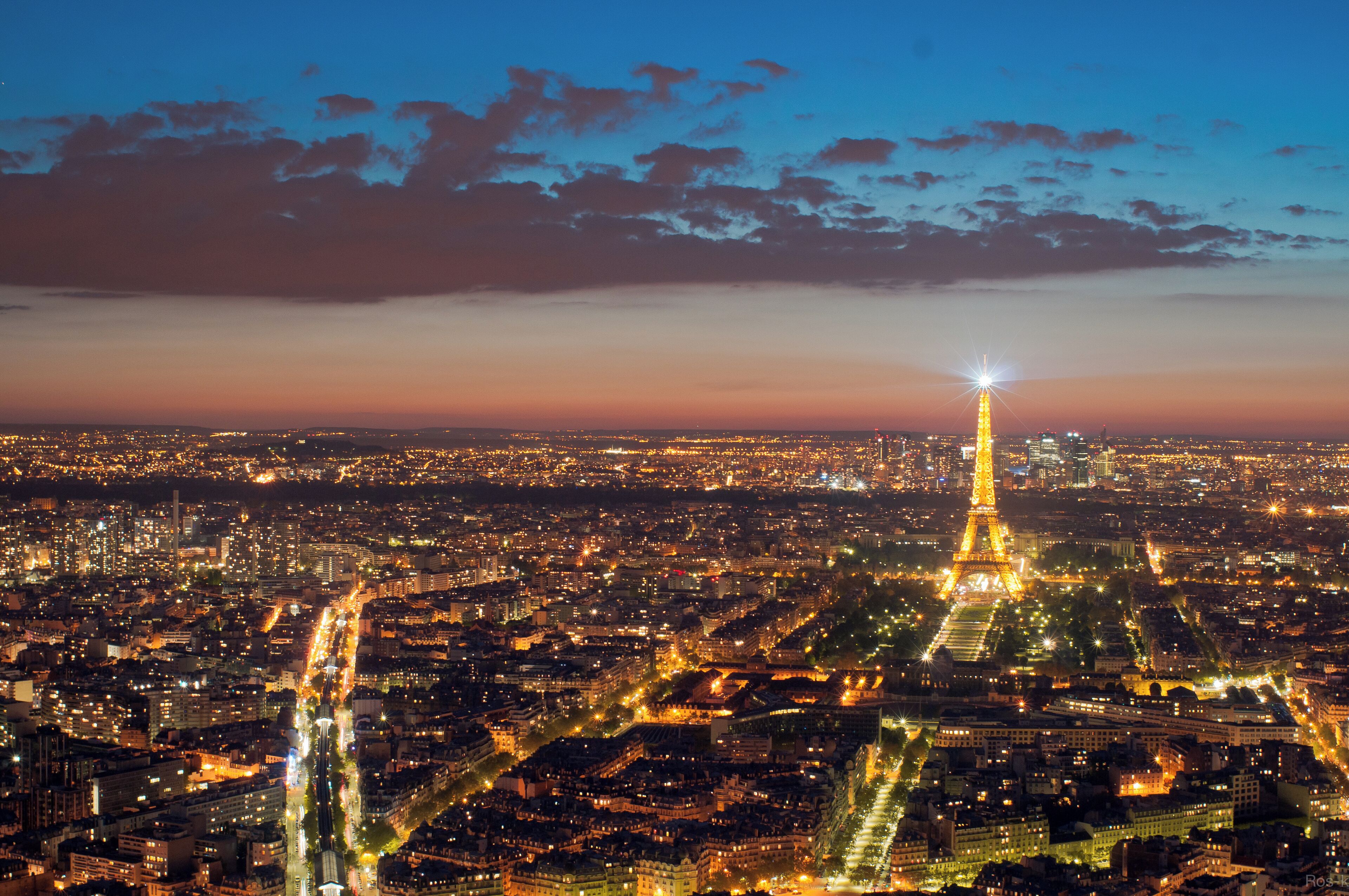 A nighttime cityscape with the Eiffel Tower illuminated, surrounded by dense urban buildings and a river.