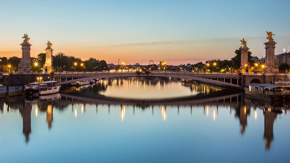 Uma ponte histórica com estátuas, um rio calmo refletindo a estrutura, e luzes da cidade ao fundo.