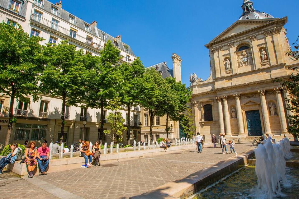 Une place historique avec une fontaine, entourée d’arbres et bordée d’édifices d’architecture classique.