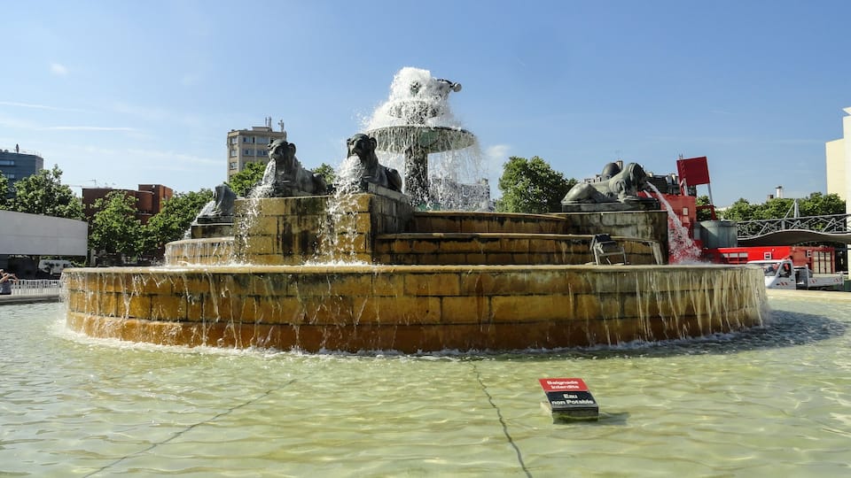 A fountain with statues and water jets, surrounded by a paved area.