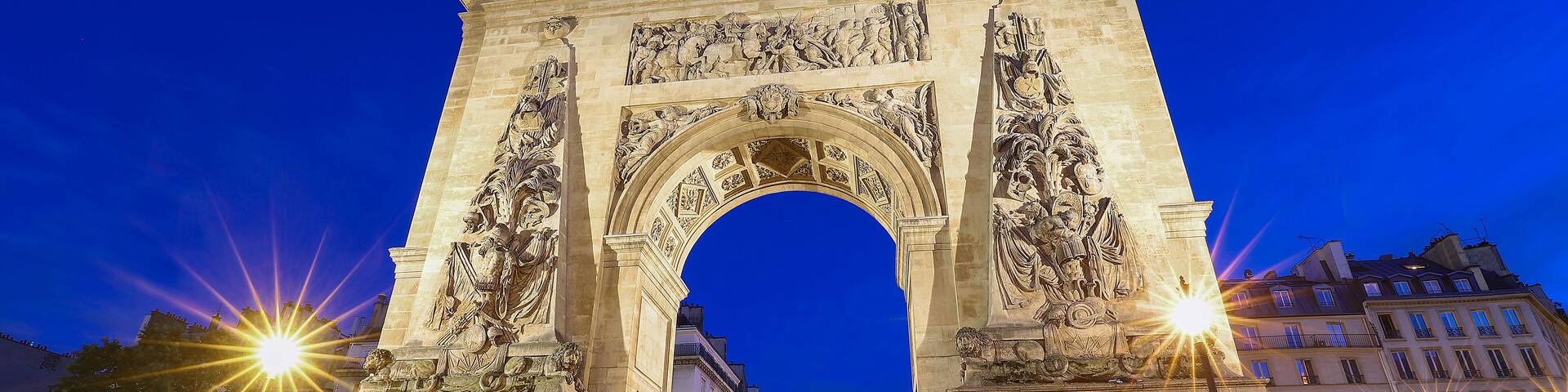 El Arco del Triunfo iluminado en la noche, con edificios circundantes y calles empedradas.