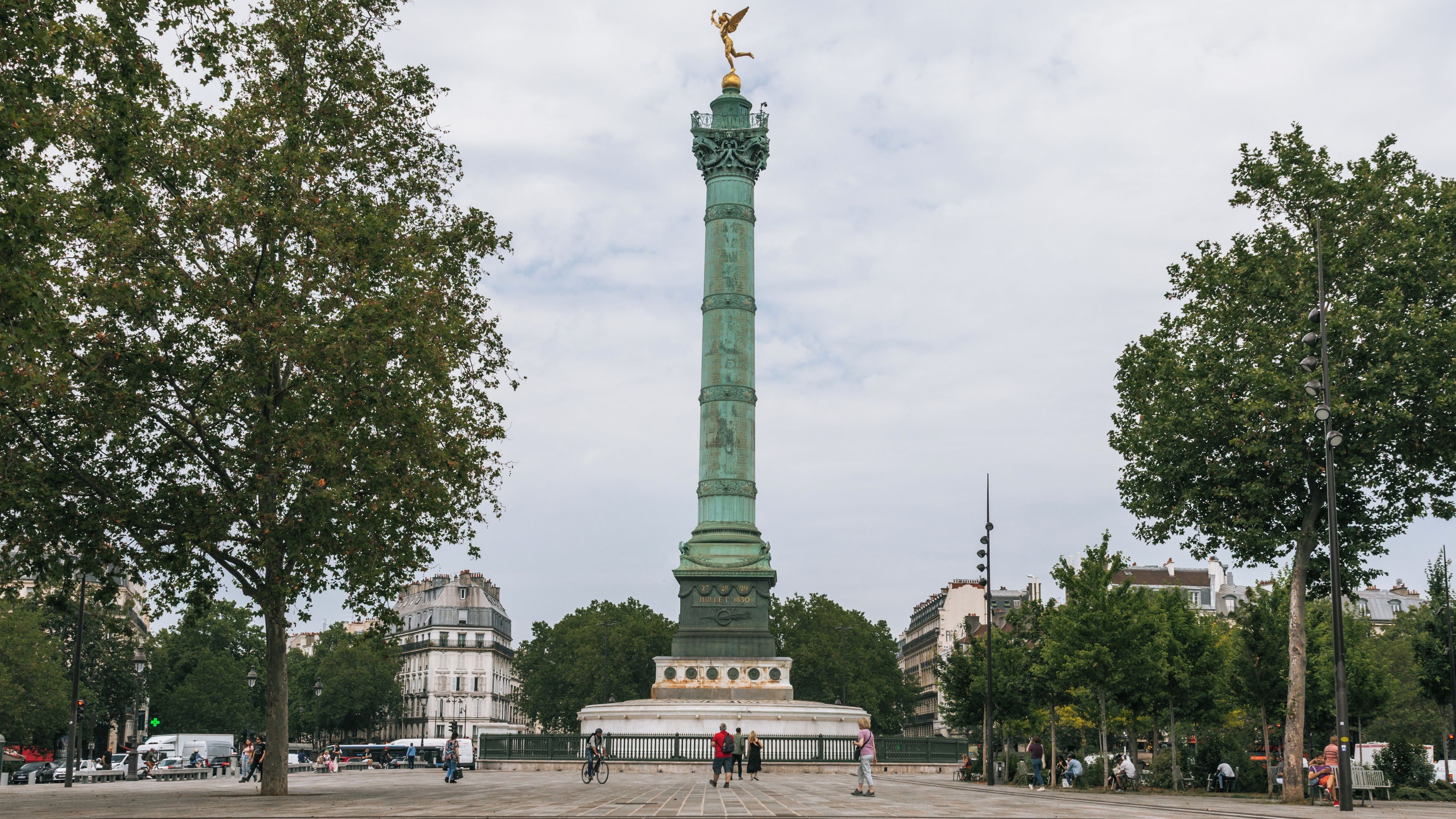 Ein hohes, grünes Denkmal mit einer goldenen Statue auf der Spitze, umgeben von Bäumen und Gebäuden.