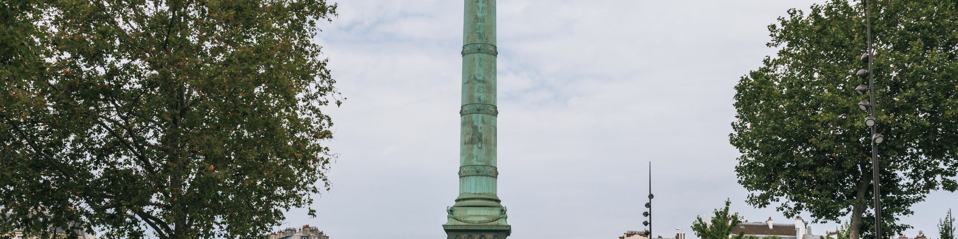 Un monumento alto de color verde con una estatua dorada en la cima, rodeado de árboles y edificios.