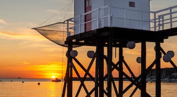 Ein weißes Gebäude mit roter Tür auf einem Steg, umgeben von Wasser und Booten im Sonnenuntergang.
