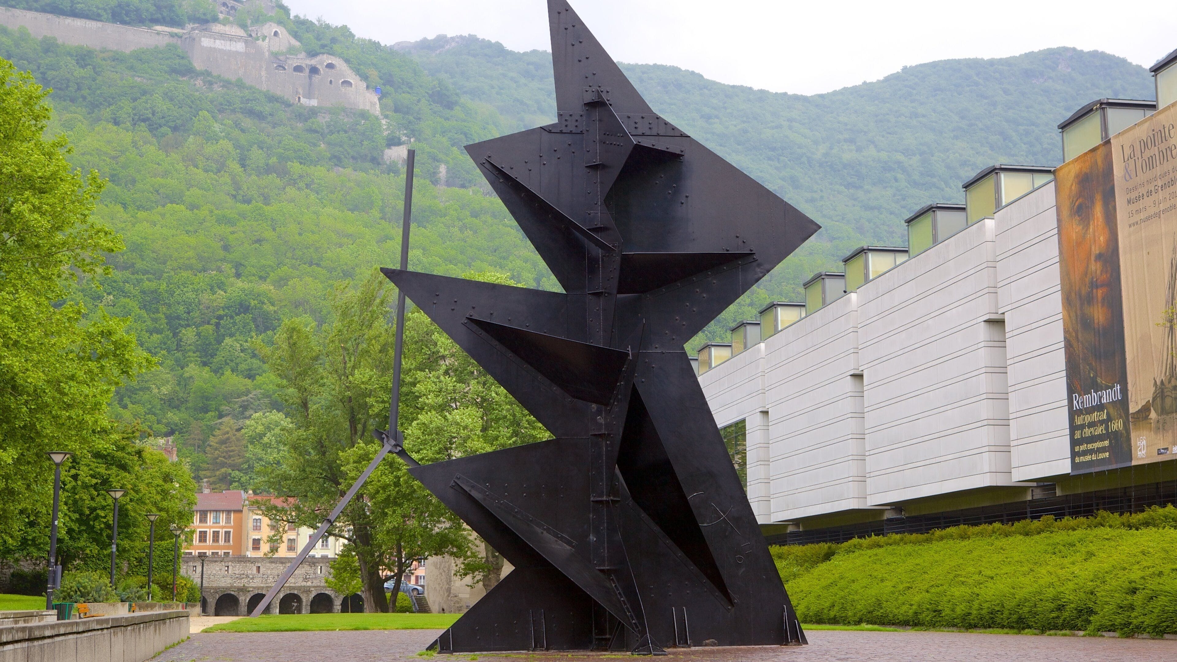 Une sculpture moderne dans un parc urbain, avec en arrière-plan des collines verdoyantes et un bâtiment historique.