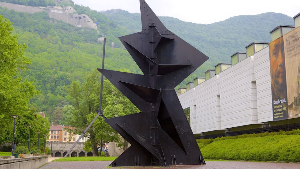 Une sculpture moderne dans un parc urbain, avec en arrière-plan des collines verdoyantes et un bâtiment historique.