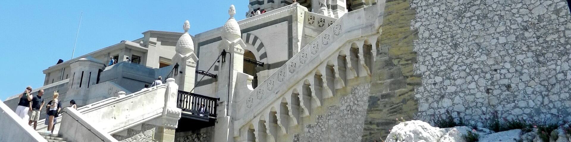A historic tower with intricate stonework and statues, a stone staircase leading up to it, and a clear blue sky in the background.
