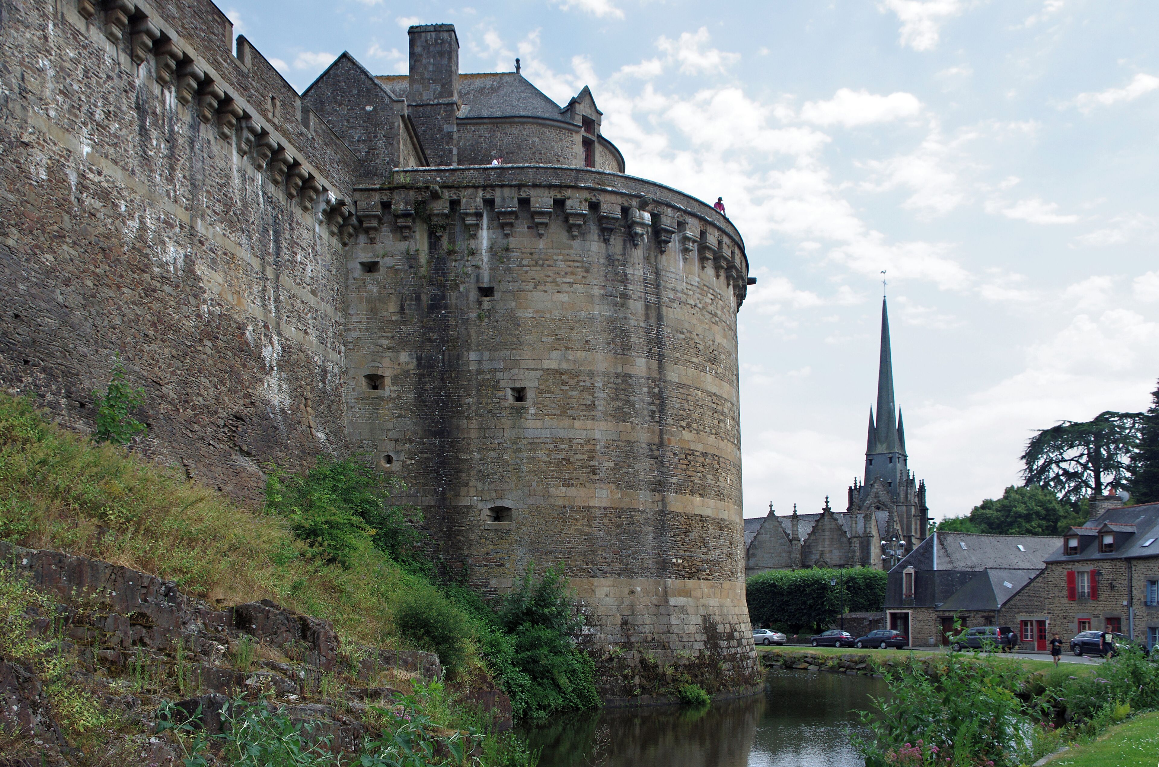 A historic stone castle with a river in front, a church with a tall spire, and a small town with buildings and cars.