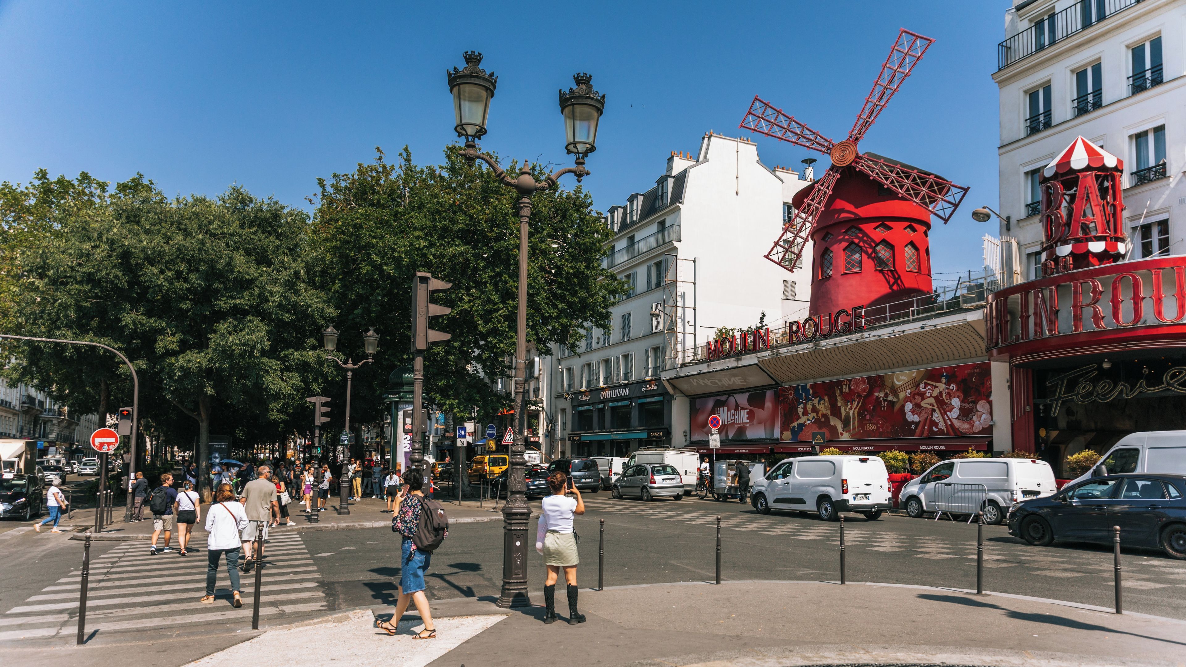 Una calle concurrida con un molino rojo destacado, un teatro antiguo y arquitectura europea clásica.